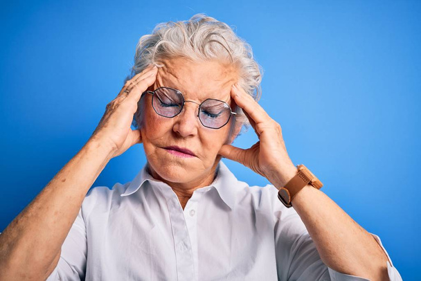 Senior kaunis nainen yllään tyylikäs paita ja lasit yli eristetty sininen tausta kärsii päänsärky epätoivoinen ja korosti, koska kipu ja migreeni. Kädet pään päälle.
. - Valokuva, kuva