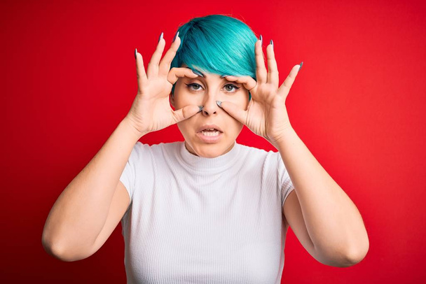Young beautiful woman with blue fashion hair wearing casual t-shirt over red background Trying to open eyes with fingers, sleepy and tired for morning fatigue - Foto, afbeelding