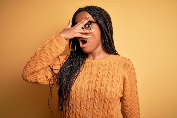 Joven afroamericana mujer inteligente con gafas y suéter casual sobre fondo amarillo asomándose en la cara cubierta de choque y los ojos con la mano, mirando a través de los dedos con expresión avergonzada
. - Foto, imagen