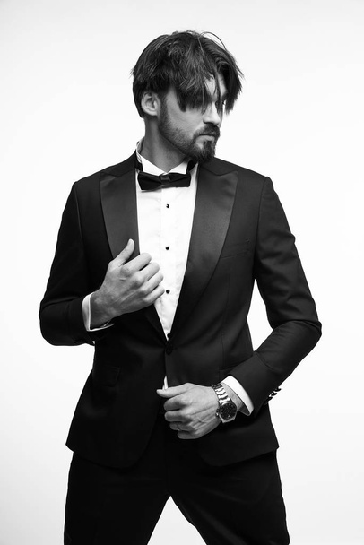black and white portrait of a handsome young man with short beard and dark hair, he is wearing a black tuxedo, a white classic shirt with a black bow tie, he touches his jacket with his hands and looks away - Valokuva, kuva