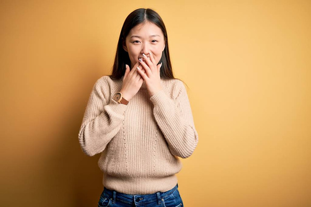 Nuori kaunis aasialainen nainen yllään rento villapaita yli keltainen eristetty tausta nauraa ja hämmentynyt kikattaa peittää suun kädet, juorut ja skandaali käsite
 - Valokuva, kuva