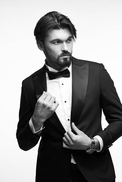 black and white portrait of a handsome young man with short beard and dark hair, he is wearing a black tuxedo, a white classic shirt with a black bow tie, he touches his jacket with his hands and looks away - Valokuva, kuva