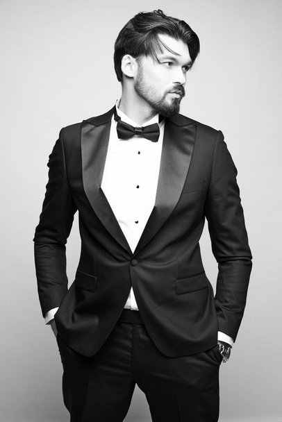 black and white portrait of a handsome young man with short beard and dark hair, he is wearing a black tuxedo, a white classic shirt with a black bow tie, he looks away, hands in pockets - Фото, зображення