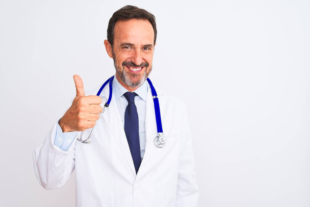 Middle age doctor man wearing coat and stethoscope standing over isolated white background doing happy thumbs up gesture with hand. Approving expression looking at the camera showing success. - Zdjęcie, obraz