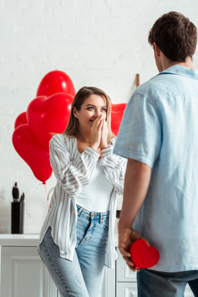 back view of man holding heart-shaped gift box near excited girlfriend on valentines day  - Photo, Image
