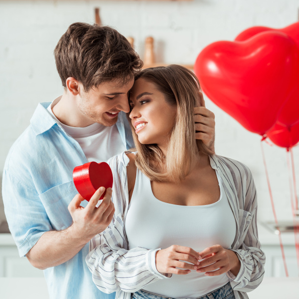 man holding heart-shaped gift box and looking at cheerful girlfriend with big breast  - Fotografie, Obrázek