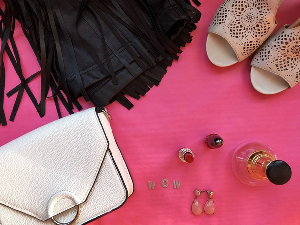 Witte schoenen, witte handtas, zwarte leren rok met franje op roze achtergrond. Parfum, rode lippenstift, rode nagellak, oorbellen. Stijlvol plat. Vrouwelijke mode. Kopieerruimte  - Foto, afbeelding