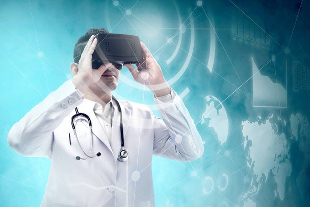 Asian male doctor in white coat and stethoscope using virtual reality device and looking at virtual screen which display the interface of financial chart and world maps. Digital technology concept - Fotografie, Obrázek