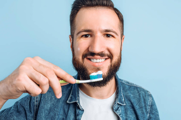 A young handsome man with a beard is brushing his teeth on an isolated blue background with raised - Fotoğraf, Görsel