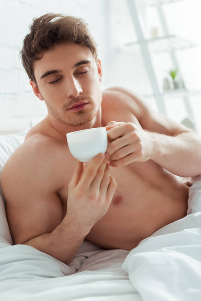 félmeztelen férfi csukott szemmel kezében csésze kávét az ágyban  - Fotó, kép
