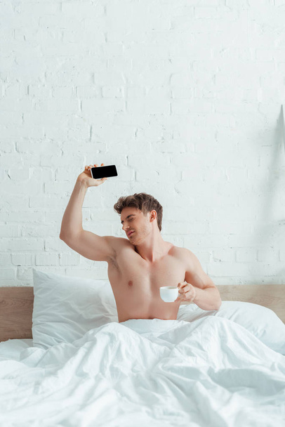 ベッドの上でコーヒーとスマートフォンを持ってるみすぼらしい男  - 写真・画像