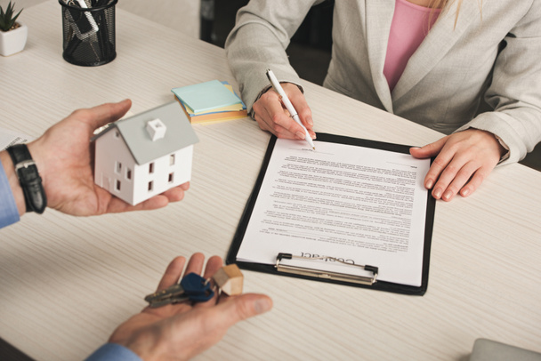 cropped view of man holding keys and house model near woman signing contract  - Photo, Image