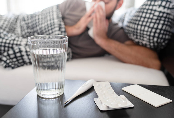 medication, thermometer and glass of water on nightstand against sick male person lying in bed - Foto, Imagem