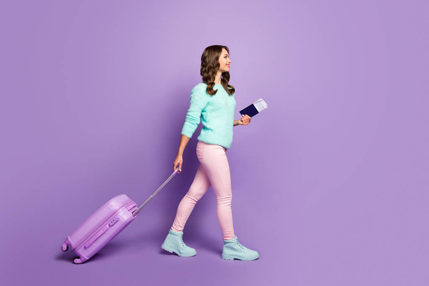 Full length photo of pretty funny lady walking airport registration with tickets rolling trip suitcase wear fuzzy sweater pink pastel trousers boots isolated purple color background - Foto, Imagen