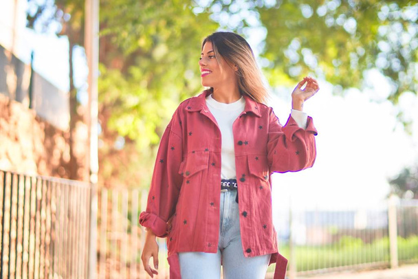 Молода красива жінка в червоній куртці посміхається щасливо і впевнено. Стоячи з посмішкою на вулиці міста
 - Фото, зображення