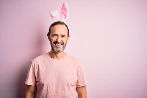 Hombre alzado de mediana edad con orejas de conejo de pie sobre un fondo rosa aislado con una sonrisa feliz y fresca en la cara. Persona afortunada
. - Foto, Imagen