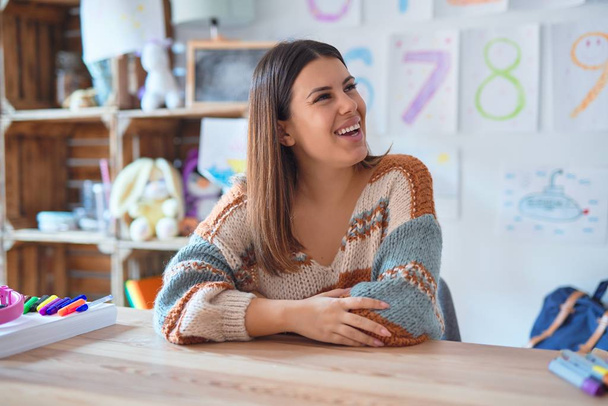 Young beautiful teacher woman wearing sweater and glasses sitting on desk at kindergarten looking away to side with smile on face, natural expression. Laughing confident. - Photo, Image