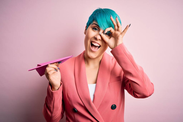 Νεαρή επιχειρηματίας με μπλε μαλλιά μόδας κρατώντας αεροπλάνο χαρτί ως ταξιδιωτικό σύμβολο με χαρούμενο πρόσωπο χαμογελώντας κάνει ok σημάδι με το χέρι στο μάτι κοιτάζοντας μέσα από τα δάχτυλα - Φωτογραφία, εικόνα
