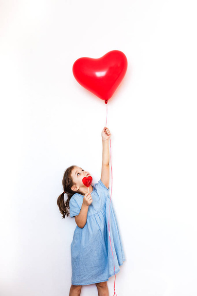 a little girl holding a beautiful red heart-shaped balloon for a Valentine's day gift and a Lollipop in the shape of a heart, lovers, Valentine's day, family and heart - Zdjęcie, obraz