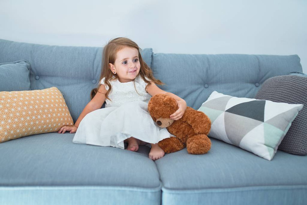 Adorable niño rubio sentado en el sofá jugando con el oso de peluche en casa
 - Foto, Imagen