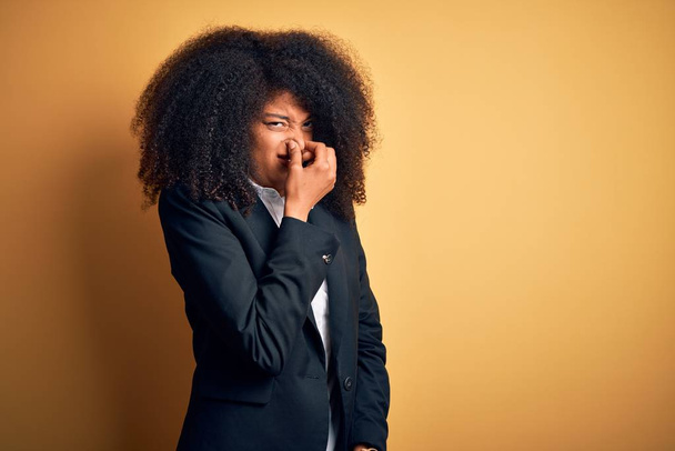 Joven mujer de negocios afroamericana hermosa con el pelo afro usando chaqueta elegante oliendo algo apestoso y repugnante, olor intolerable, conteniendo la respiración con los dedos en la nariz. Mal olor
 - Foto, imagen