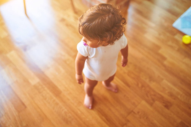 Hermosa niña pequeña con camiseta blanca de pie en el suelo usando chupete
 - Foto, Imagen