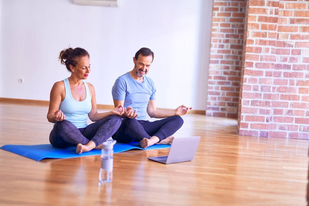 Middle age beautiful sporty couple smiling happy. Sitting on mat practicing yoga doing lotus pose using laptop at gym - Photo, Image