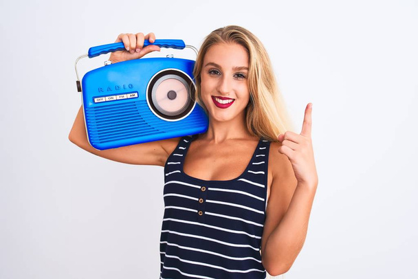 Nuori kaunis nainen, jolla on vintage-radio seisomassa eristetyn valkoisen taustan päällä yllättynyt ideasta tai kysymyksestä, joka osoittaa sormella onnellisia kasvoja, numero yksi
 - Valokuva, kuva