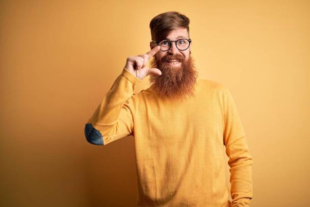 Красивый ирландский рыжий мужчина с бородой в очках на жёлтом изолированном фоне улыбается и уверенно жестикулирует рукой, делая знак небольшого размера с пальцами и камерой. Концепция измерения
. - Фото, изображение