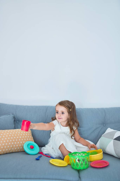Αξιολάτρευτο ξανθό νήπιο κάθεται στον καναπέ μαγείρεμα με τα τρόφιμα και μαχαιροπήρουνα παιχνίδια στο σπίτι - Φωτογραφία, εικόνα