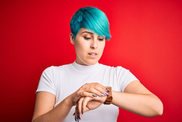 Joven hermosa mujer con el pelo de moda azul con camiseta casual sobre fondo rojo Comprobación de la hora en el reloj de pulsera, relajado y seguro
 - Foto, imagen