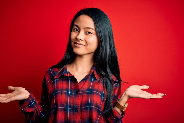 Fiatal gyönyörű kínai nő visel alkalmi ing felett elszigetelt piros háttér mosoly mutatja mindkét kezét nyitott tenyér, bemutató és reklám összehasonlítás és az egyensúly - Fotó, kép