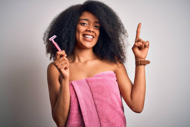 Mladá africká Američanka s afro vlasy v ručníku drží ženskou břitvu pro depilaci těla překvapena nápadem nebo otázkou ukazující prstem se šťastnou tváří, číslo jedna - Fotografie, Obrázek