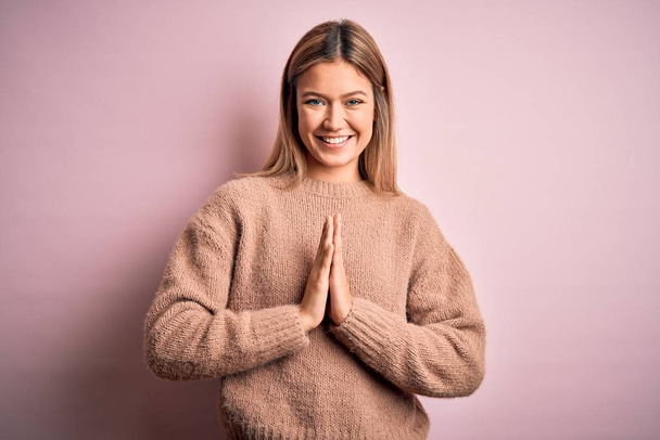 Młoda piękna blondynka ubrana w zimowy wełniany sweter na różowym tle modląc się dłońmi razem prosząc o przebaczenie uśmiechając się pewnie. - Zdjęcie, obraz