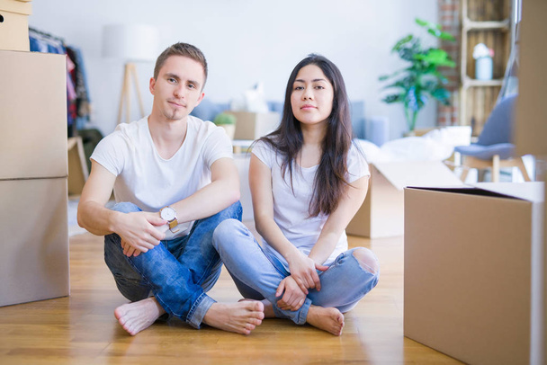 Νεαρό όμορφο ζευγάρι κάθεται στο πάτωμα στο νέο σπίτι γύρω από κουτιά από χαρτόνι με σοβαρή έκφραση στο πρόσωπο. Απλή και φυσική κοιτάζοντας την κάμερα. - Φωτογραφία, εικόνα
