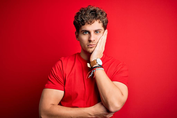 Young blond handsome man with curly hair wearing casual t-shirt over red background thinking looking tired and bored with depression problems with crossed arms. - Photo, Image