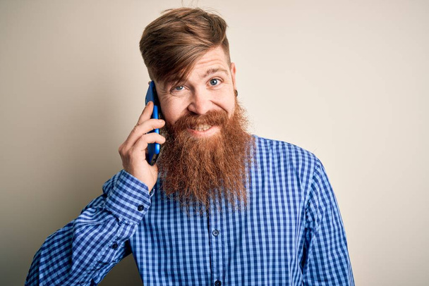Pelirrojo irlandés con barba conversando en un teléfono inteligente sobre un fondo aislado con una cara feliz de pie y sonriendo con una sonrisa confiada mostrando los dientes
 - Foto, imagen