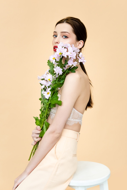 beautiful woman holding blooming flowers and sitting on chair isolated on beige  - 写真・画像