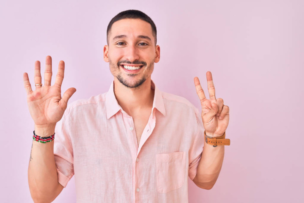 junger gutaussehender Mann in rosa Hemd, der vor isoliertem Hintergrund steht und mit Finger Nummer sieben nach oben zeigt, während er selbstbewusst und glücklich lächelt. - Foto, Bild