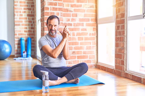 Esportista de meia idade bonito sentado no tapete fazendo alongamento exercício de ioga no ginásio Segurando arma simbólica com gesto de mão, jogando matar armas de tiro, rosto com raiva
 - Foto, Imagem