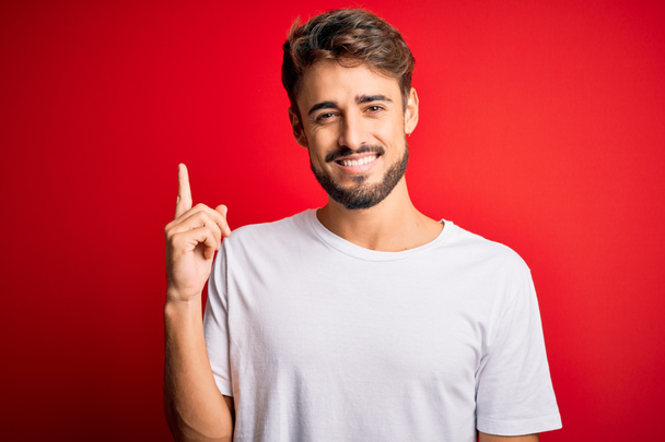 Νεαρός όμορφος άντρας με γενειάδα φορώντας casual t-shirt πάνω από κόκκινο φόντο με ένα μεγάλο χαμόγελο στο πρόσωπο, δείχνοντας με το χέρι και το δάχτυλο στο πλάι κοιτάζοντας την κάμερα. - Φωτογραφία, εικόνα