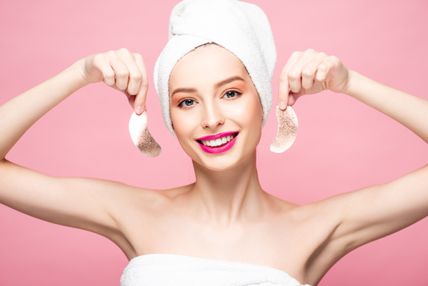 χαμογελαστή νεαρή γυναίκα με πετσέτα κρατώντας μπαλώματα ματιών απομονώνονται σε ροζ  - Φωτογραφία, εικόνα