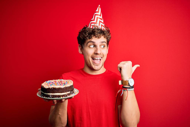 Young blond man with curly hair wearing birthday hat holding cake over red background pointing and showing with thumb up to the side with happy face smiling - Photo, Image