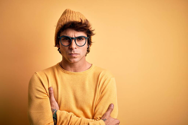 Young handsome man wearing casual t-shirt and glasses over isolated yellow background skeptic and nervous, disapproving expression on face with crossed arms. Negative person. - Fotoğraf, Görsel