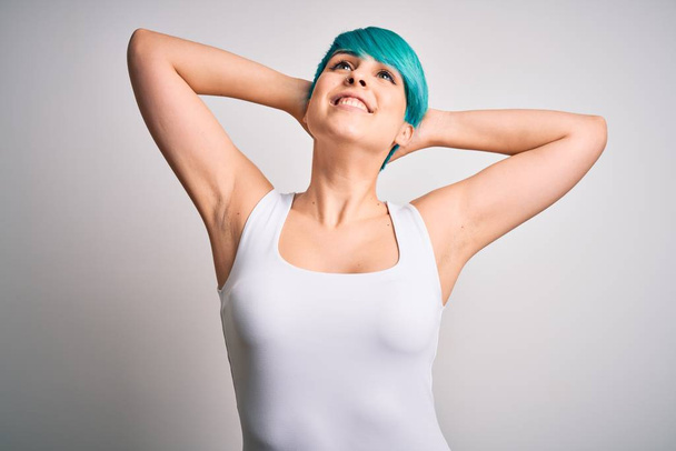 Mavi saçlı genç bir kadın beyaz arka plan üzerine günlük tişört giyip rahatlıyor ve esniyor, kolları ve elleri başının ve boynunun arkasında mutlu bir şekilde gülümsüyor. - Fotoğraf, Görsel