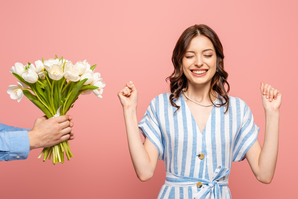 cropped view of man presenting bouquet of white tulips to excited young woman showing winner gesture isolated on pink - Fotó, kép