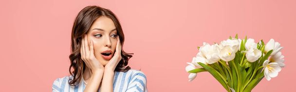 panoramic shot of surprised girl touching face while looking at bouquet of white tulips isolated on pink - Foto, afbeelding
