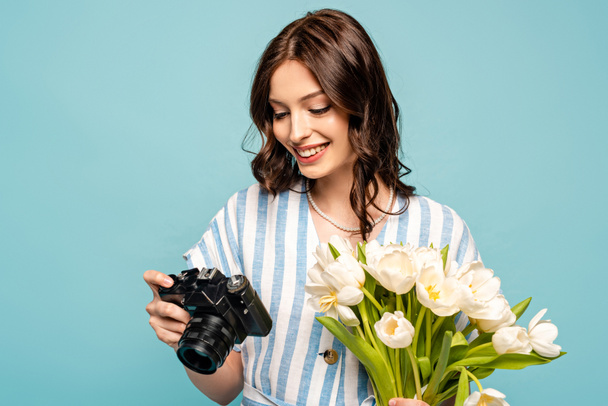 cheerful young woman holding digital camera and bouquet of white tulips isolated on blue - Фото, изображение