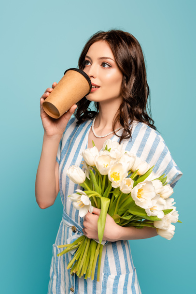 snivá mladá žena pije kávu jít, zatímco drží bílé tulipány izolované na modré - Fotografie, Obrázek