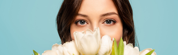 panoramic shot of young woman enjoying flavor of white tulips while looking at camera isolated on blue - Foto, Bild